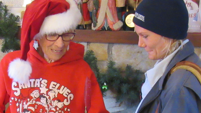 A USPS letter carrier delivers a package to a woman wearing a Santa Claus cap and red hooded sweatshirt