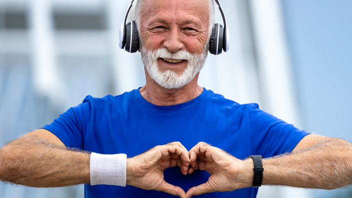 A white-haired man in a blue shirt makes a heart symbol with his hands while wearing exercise gear and standing on a bridge