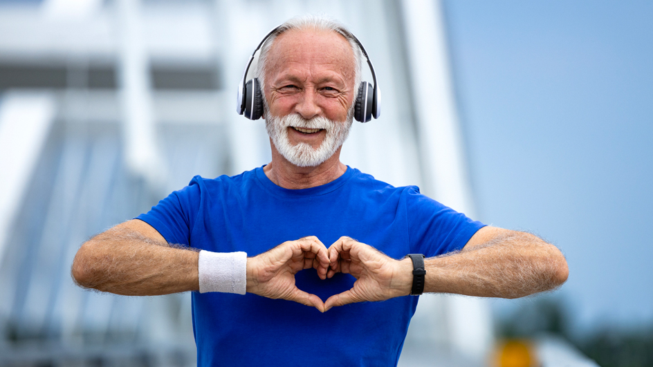 A white-haired man in a blue shirt makes a heart symbol with his hands while wearing exercise gear and standing on a bridge