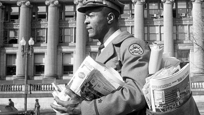 A black-and-white image of a Black letter carrier wearing a postal uniform and carrying a mail satchel in front of a historic Post Office building