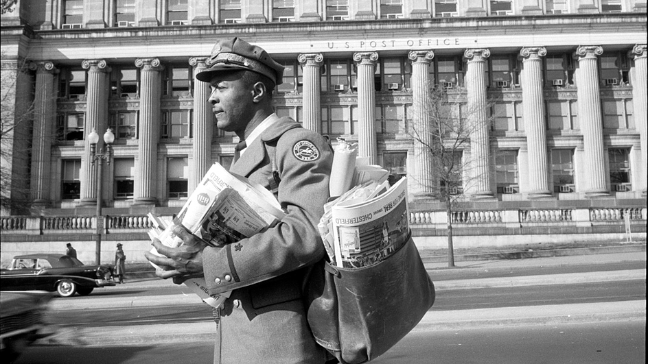 A black-and-white image of a Black letter carrier wearing a postal uniform and carrying a mail satchel in front of a historic Post Office building