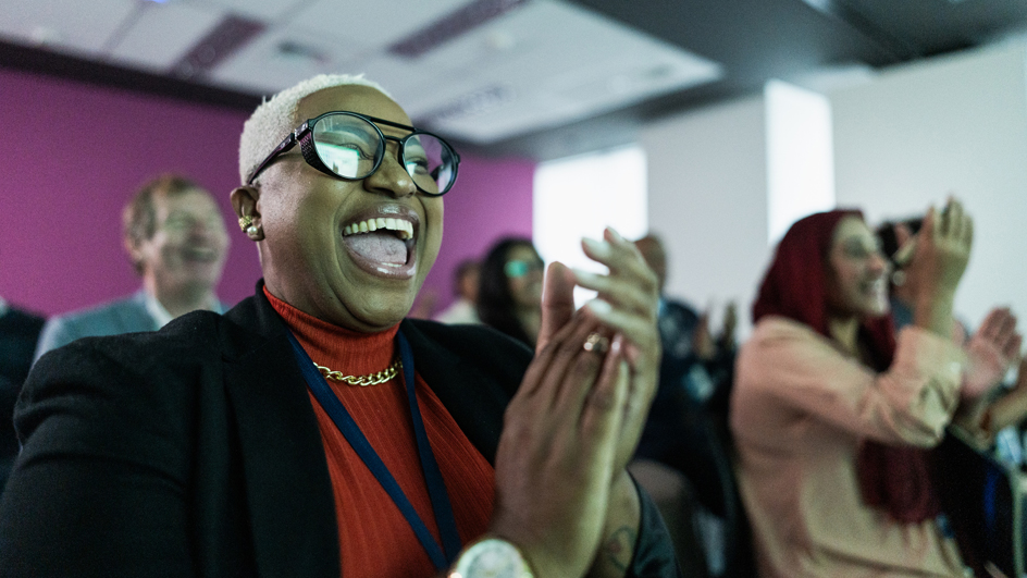 A woman smiles and claps while seated in an auditorium. She is surrounded by other people who are cheerfully applauding.