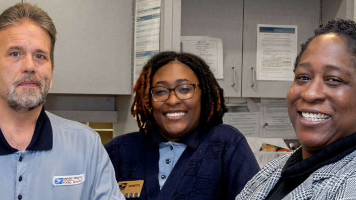 Two people in postal uniforms stand behind the retail counter in a Post Office while a woman in business attire stands in front