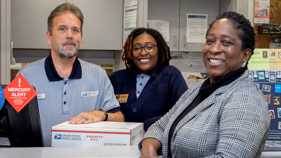 Two people in postal uniforms stand behind the retail counter in a Post Office while a woman in business attire stands in front