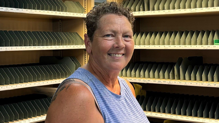 A woman in a light blue shirt stands in front of a letter-sorting rack.