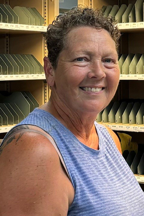 A woman in a light blue shirt stands in front of a letter-sorting rack.