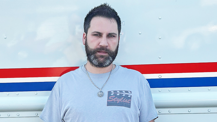 A man stands next to a USPS delivery vehicle. He wears a T-shirt and has a silver chain with a medallion around his neck.