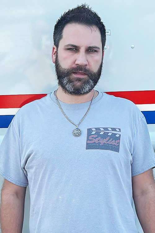 A man stands next to a USPS delivery vehicle. He wears a T-shirt and has a silver chain with a medallion around his neck.