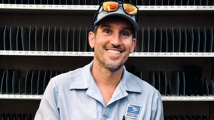 A man wearing a USPS uniform shirt stands in a Post Office workroom, smiling