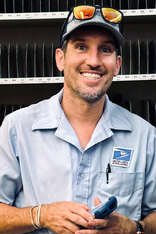 A man wearing a USPS uniform shirt stands in a Post Office workroom, smiling