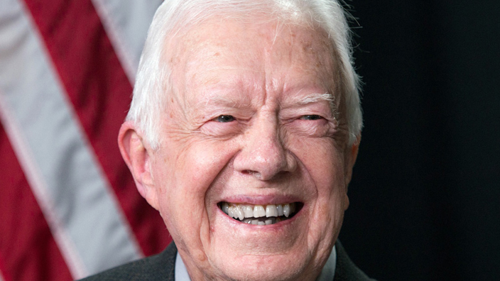 A smiling Jimmy Carter sits on a stage with a U.S. flag behind him