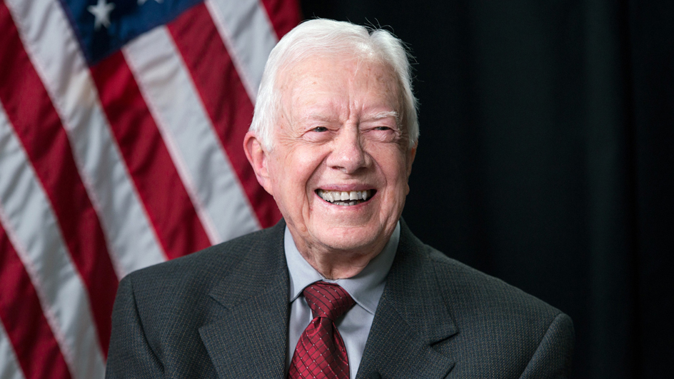 A smiling Jimmy Carter sits on a stage with a U.S. flag behind him