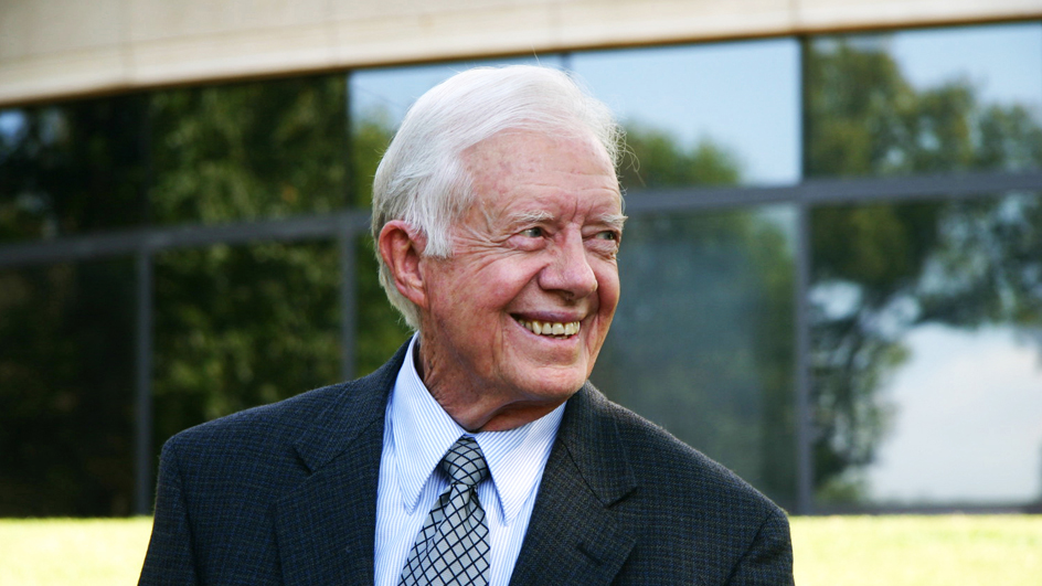 A tight close-up of a smiling Jimmy Carter, who stands in front of a glass building