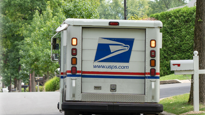 A USPS delivery vehicle is stopped on a lawn-lined street.