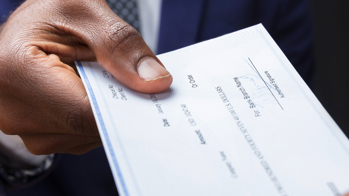 A check is being passed across a table from the hand of a man wearing a suit and tie into another person’s hand.