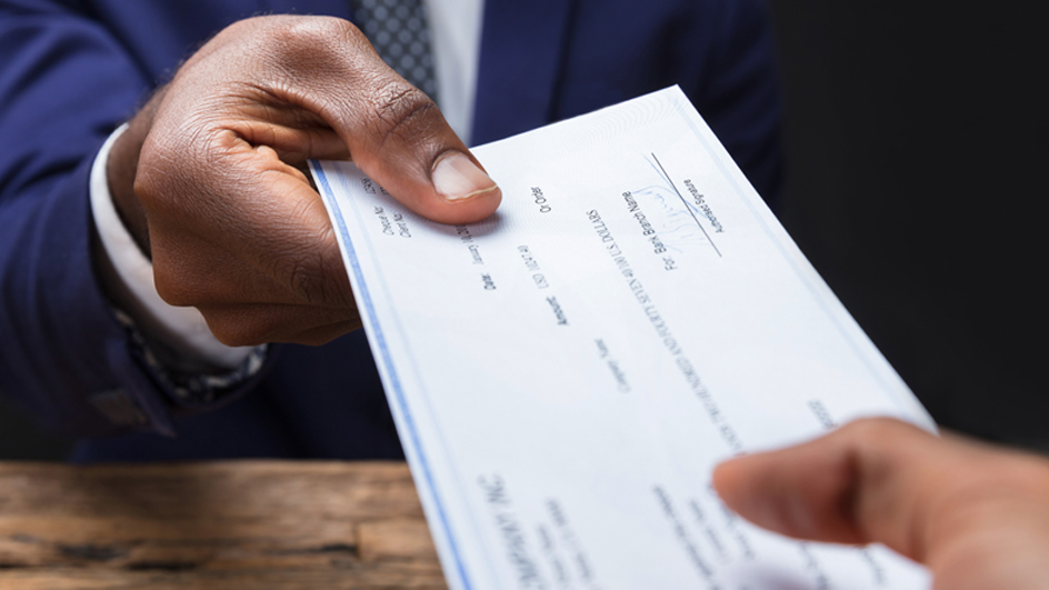 A check is being passed across a table from the hand of a man wearing a suit and tie into another person’s hand.