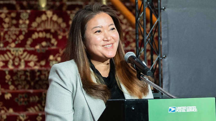 A woman in a business suit smiles while standing at a lectern. A poster that shows a postage stamp with a snake image looms behind her.