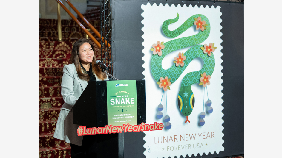 A woman in a business suit smiles while standing at a lectern. A poster that shows a postage stamp with a snake image looms behind her.