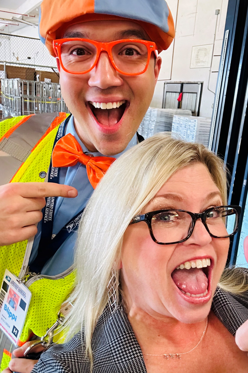 A man dressed in colorful clothing stands next to a woman in business wear. They both smile broadly.