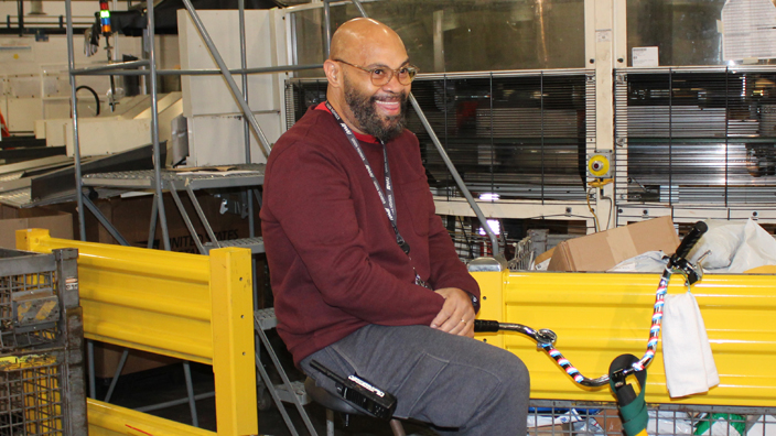 A man smiles while riding a tricycle through the workroom floor of a postal facility