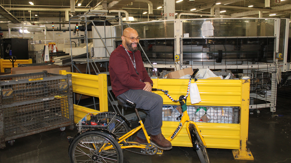 A man smiles while riding a tricycle through the workroom floor of a postal facility