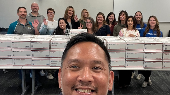 A selfie photo that shows a man in the foreground, in sharp focus, while a group of people stand behind stacks of Priority Mail boxes in the background