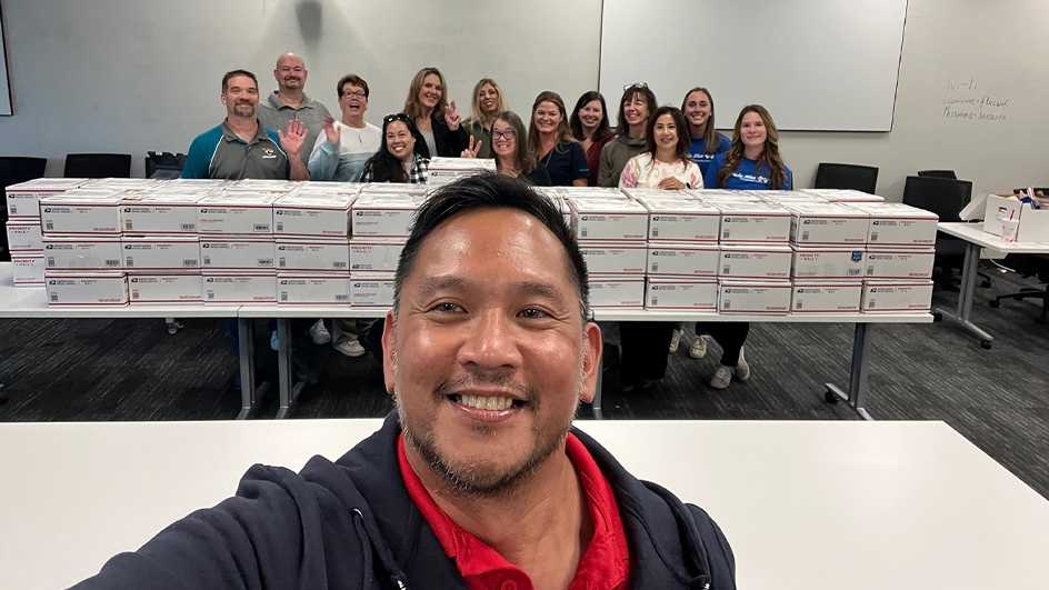 A selfie photo that shows a man in the foreground, in sharp focus, while a group of people stand behind stacks of Priority Mail boxes in the background