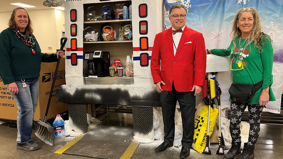 Three people stand in a postal workroom near a cardboard display that resembles the back of a postal truck, stocked with a coffee machine and winter safety items.