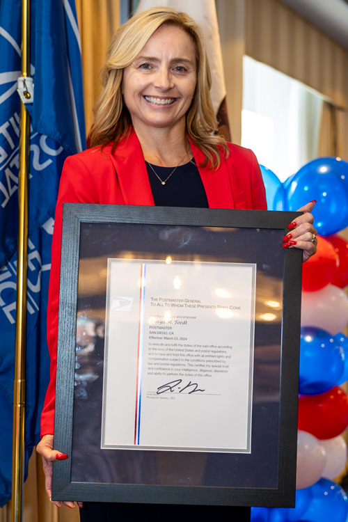 A smiling woman, dressed in a red business suit, stands on a stage with a USPS flag and balloons behind her and holds a plaque