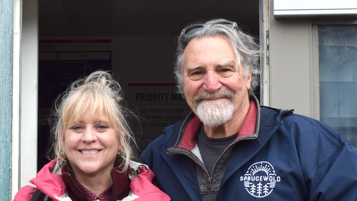 A man and woman stand in front of the door to a shingle-clad Post Office in Maine.