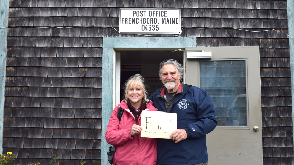 A man and woman stand in front of the door to a shingle-clad Post Office in Maine.