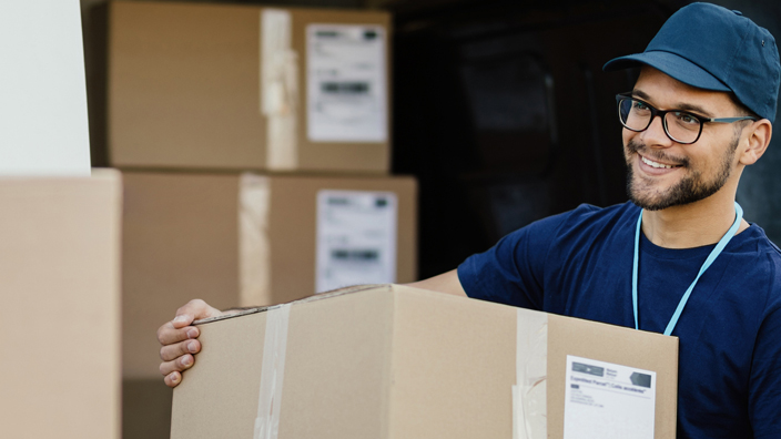 A young man wearing a blue shirt and baseball cap prepares to set a large box on a stack boxes in a warehouse setting