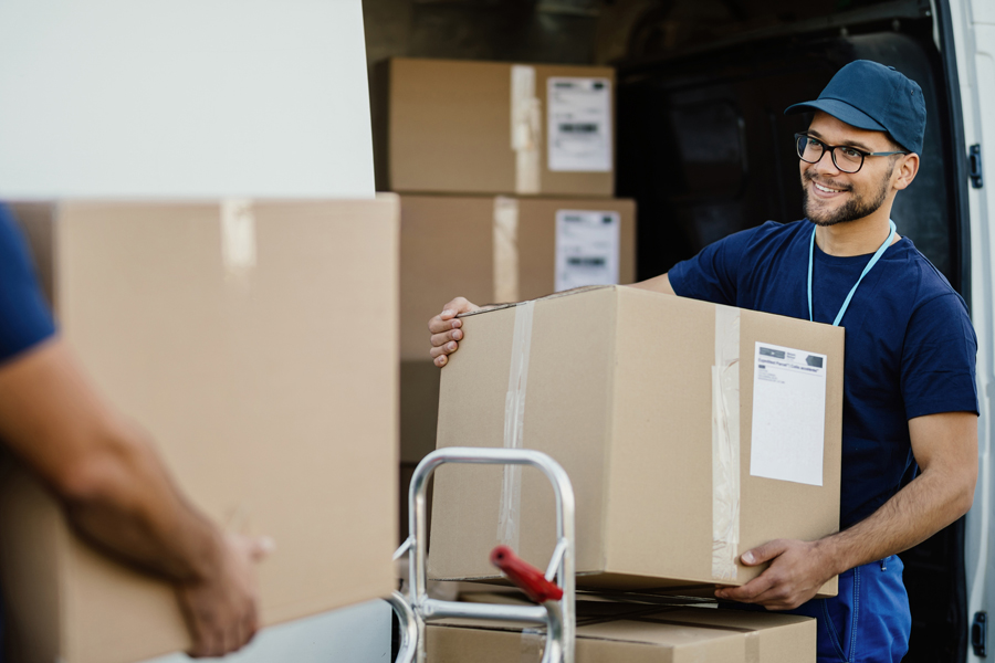 A young man wearing a blue shirt and baseball cap prepares to set a large box on a stack boxes in a warehouse setting