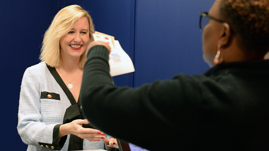 A USPS retail associate, seen from behind, hands a book of stamps to a smiling woman