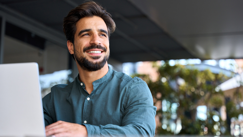 A man smiles while looking away from a laptop computer, seated in an outdoor space.