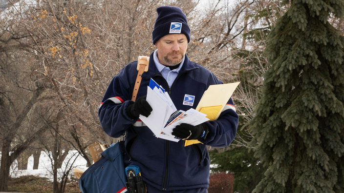 A USPS letter carrier walks along a sidewalk with snow on either side as a mountain looms in the background