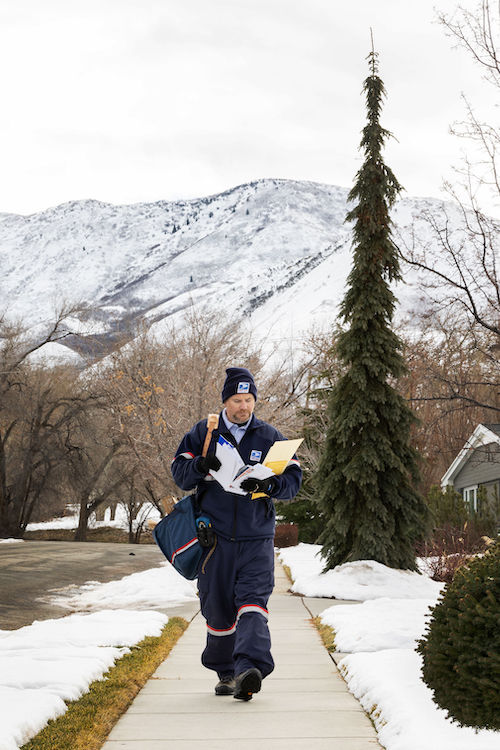 A USPS letter carrier walks along a sidewalk with snow on either side as a mountain looms in the background