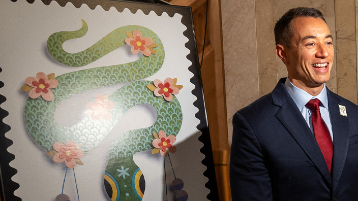 A man in a business suit stands next to a Year of the Dragon stamp poster and speaks to a video crew