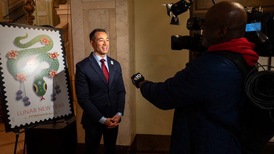 A man in a business suit stands next to a Year of the Dragon stamp poster and speaks to a video crew