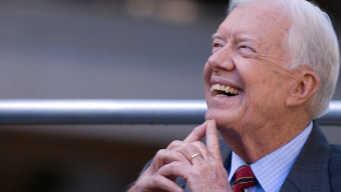 Jimmy Carter smiles brightly and looks up, resting his chin on his two index fingers, which are extended as he clasps his hands together.