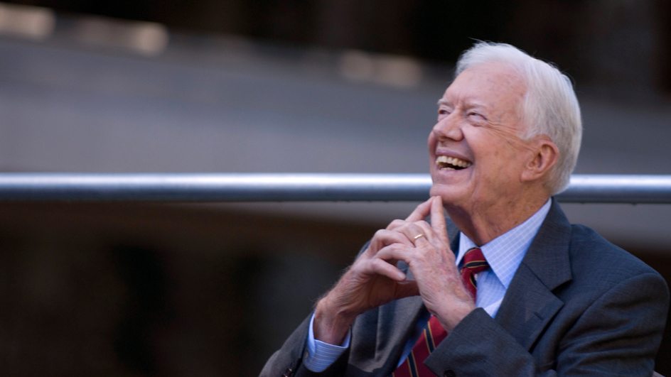 Jimmy Carter smiles brightly and looks up, resting his chin on his two index fingers, which are extended as he clasps his hands together.