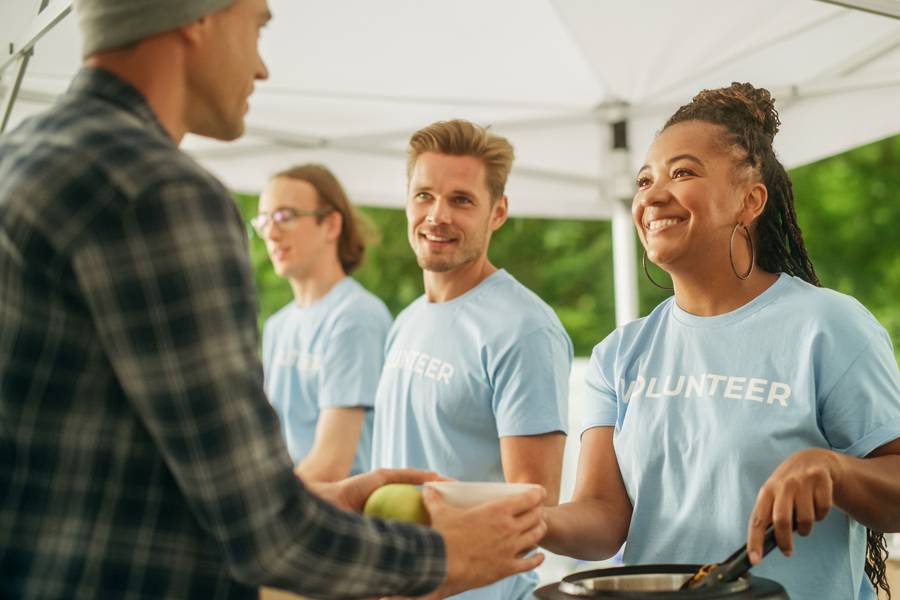 A smiling woman wearing a T-shirt that says “volunteer” dishes out food to an unhoused person