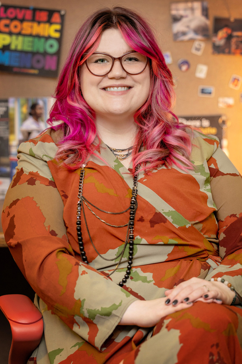 A woman with dyed purple hair, wearing a colorful dress, sits in a chair and smiles