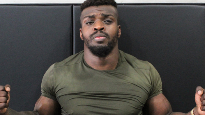 A muscular man wearing a tight shirt flexes his arms while standing along a gymnasium wall