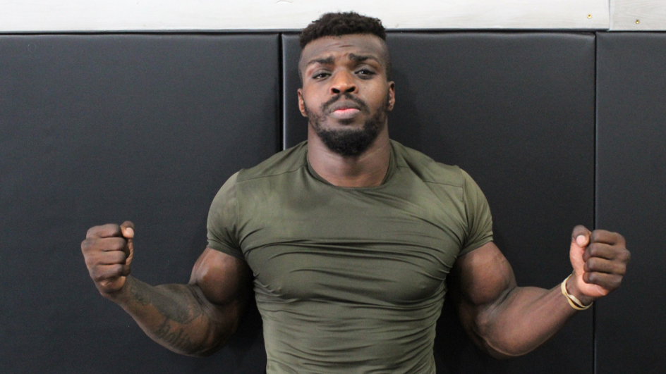 A muscular man wearing a tight shirt flexes his arms while standing along a gymnasium wall