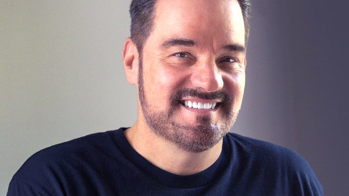 A man, seen from the chest up, sits at a keyboard and smiles. He wears a blue T-shirt with a logo that reads “Boom Box.”