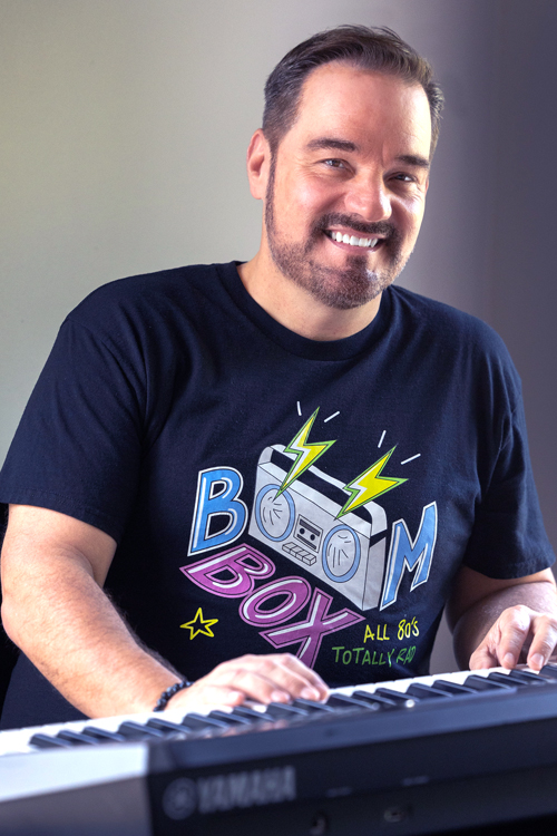 A man, seen from the chest up, sits at a keyboard and smiles. He wears a blue T-shirt with a logo that reads “Boom Box.”