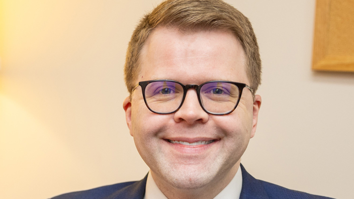 A man wearing a blue business suit and a pink tie smiles brightly