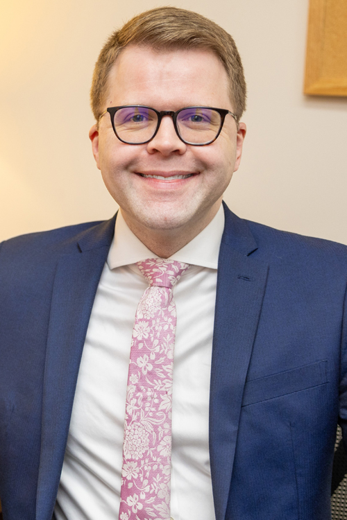 A man wearing a blue business suit and a pink tie smiles brightly