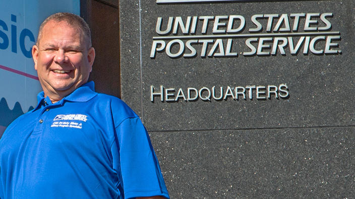 A man wearing a blue polo shirt with the USPS logo stands next to the USPS headquarters building sign.